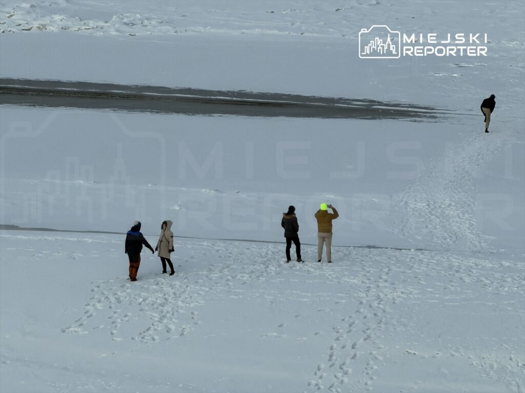 Para trzymająca się za ręce spaceruje po zaśnieżonym terenie, obok stoją dwie osoby, jedna z telefonem.