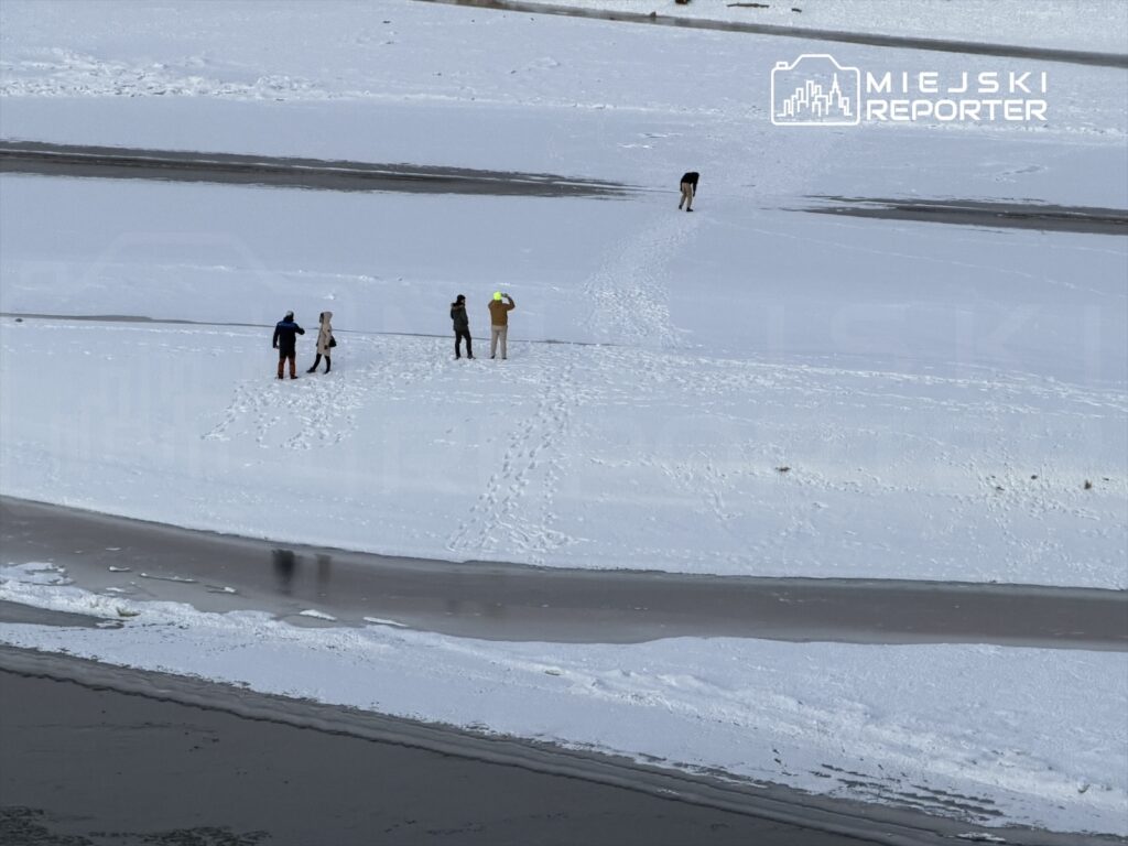 Grupa pięciu osób spaceruje po zaśnieżonym brzegu rzeki, obok płynącej wody.