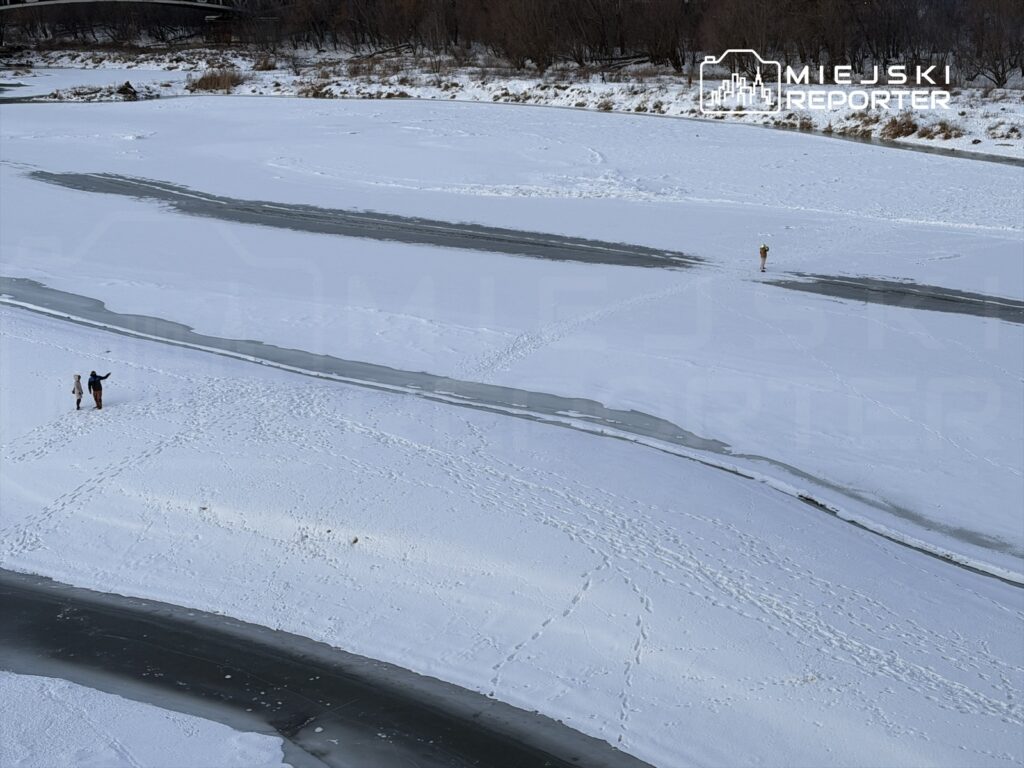 Dwie osoby spacerują po zamarzniętej rzece w zimowym krajobrazie, otoczonej śniegiem i nagimi drzewami.