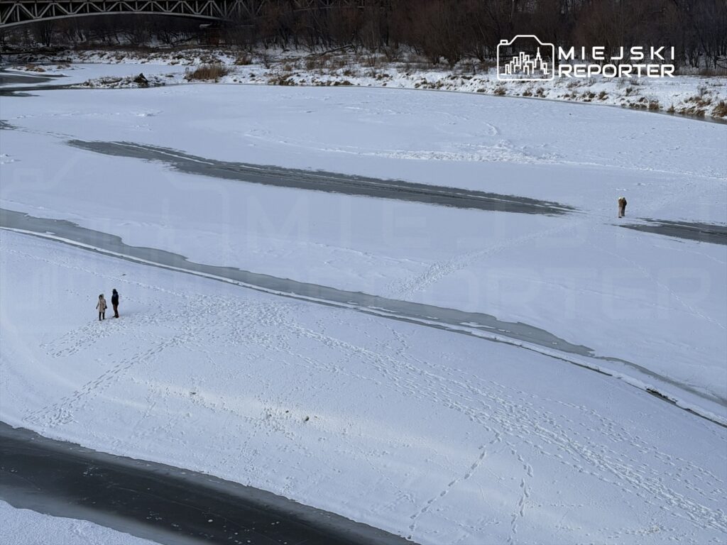 Para spacerująca po zamarzniętej rzece, w tle widoczny most i zalesione brzegi.