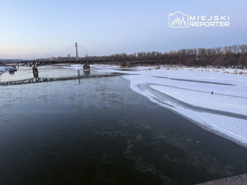Niska woda w rzece z fragmentami lodu, a w tle widać most i pojedynczą osobę spacerującą po brzegu pokrytym śniegiem.