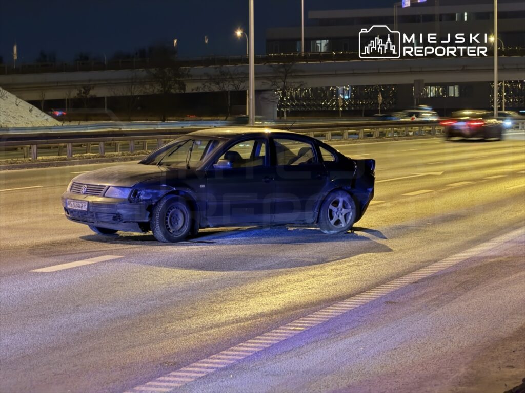 Czarny samochód osobowy stoi na oświetlonej drodze, w tle widoczne są mijające pojazdy.