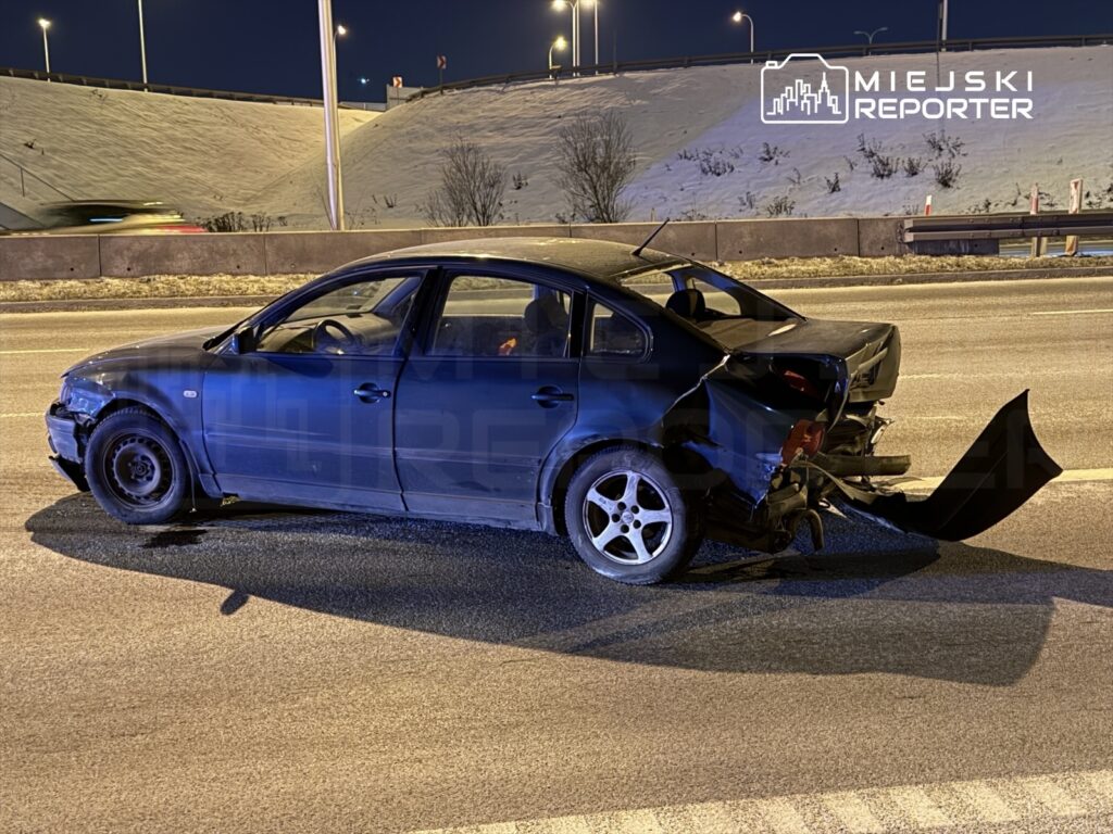 Uszkodzony czarny samochód osobowy stoi na jezdni, z widocznym wgnieceniem tylnej części i odłamanym zderzakiem.