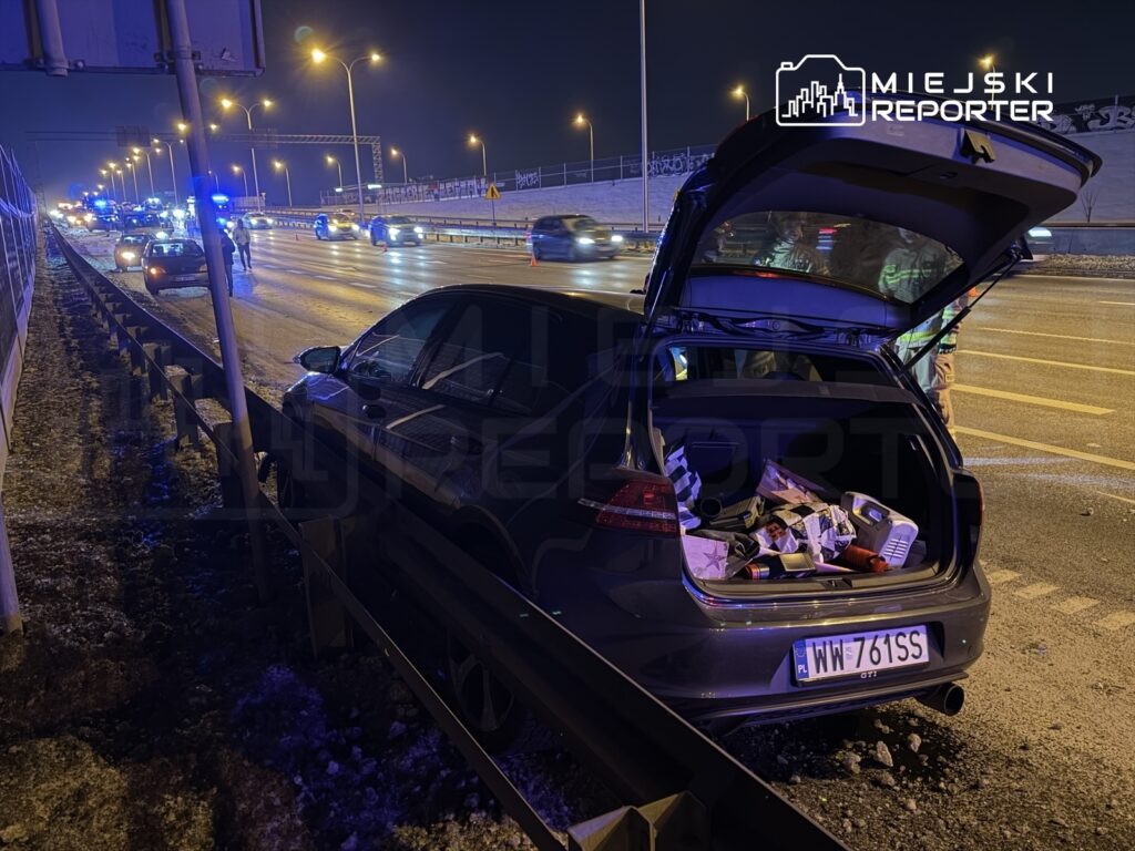 Czarny samochód zaparkowany przy drodze, z otwartą klapą bagażnika, obok stojący mężczyzna w mundurze.