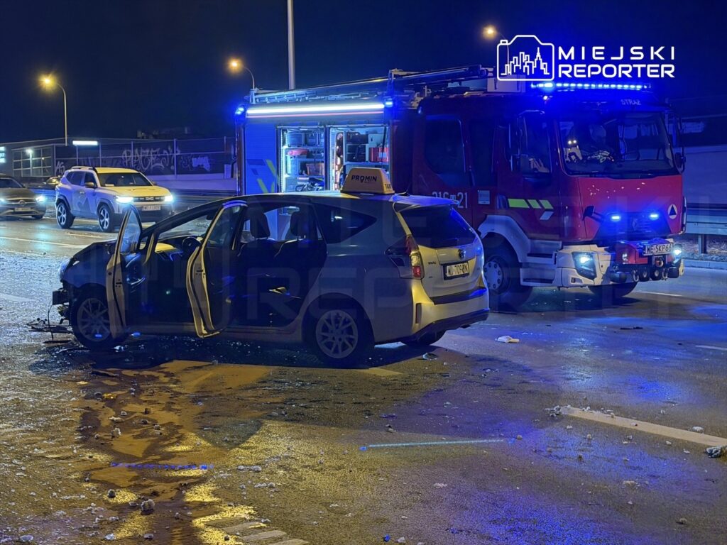 Uszkodzony samochód osobowy z otwartymi drzwiami stoi na jezdni, obok niego widać wóz strażacki z włączonymi syrenami.