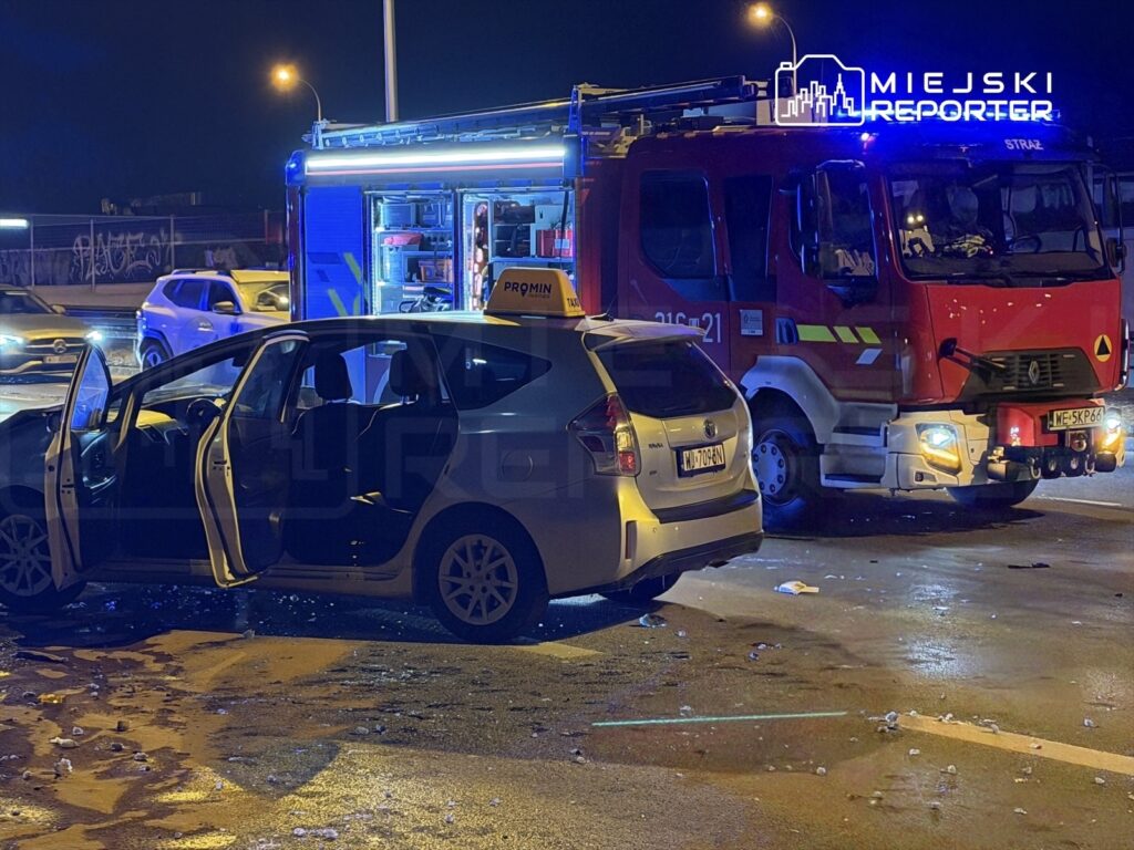 Na drodze stoi uszkodzony samochód osobowy z otwartymi drzwiami, obok zaparkowany wóz strażacki z włączonymi sygnałami świetlnymi.