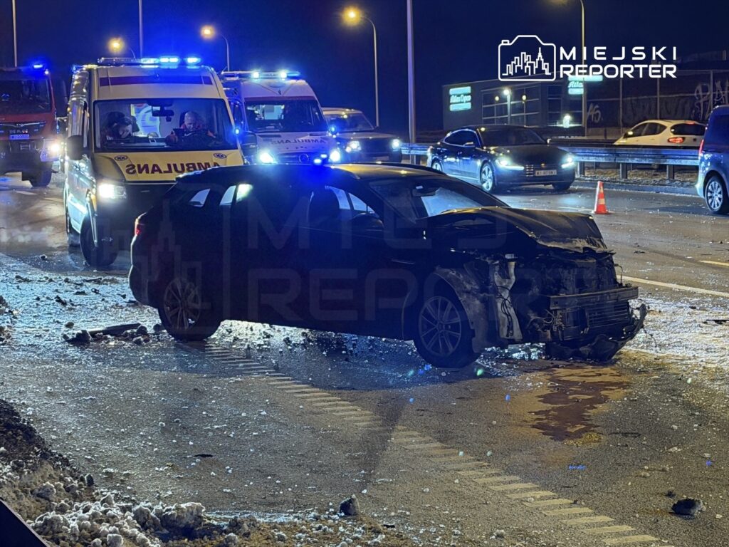 Czarny samochód osobowy z uszkodzoną przednią częścią stoi na drodze, obok niego kilka radiowozów i karetka.