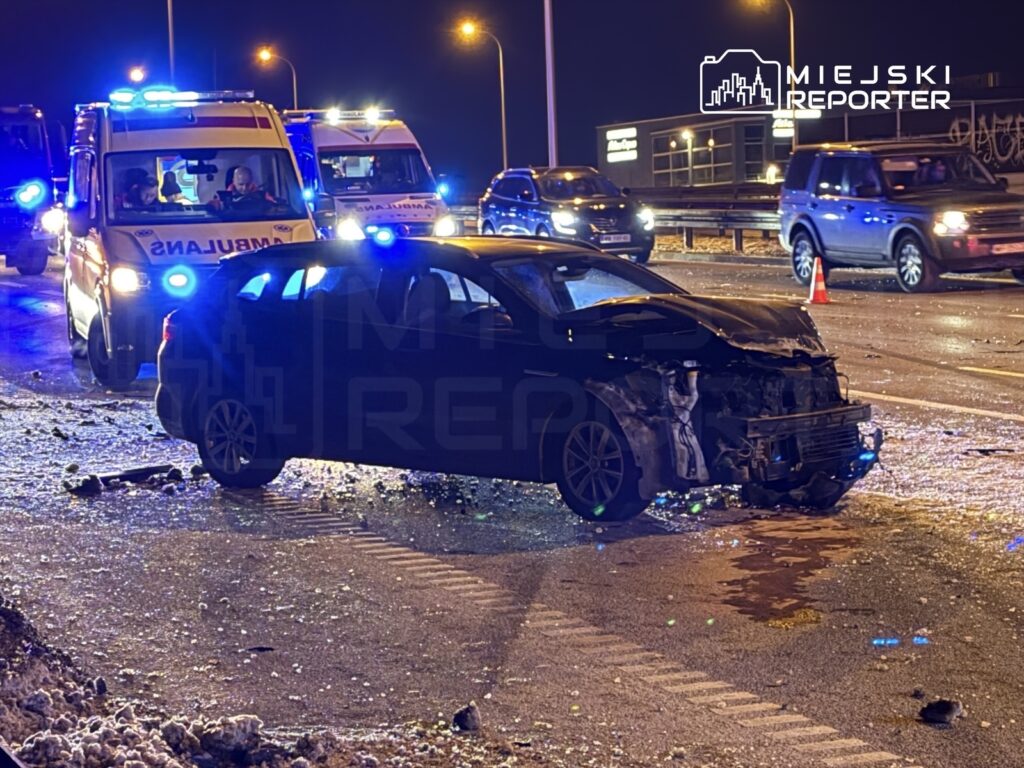 Zniszczony samochód osobowy na jezdni, obok pojazdy ratunkowe z włączonymi sygnałami świetlnymi.