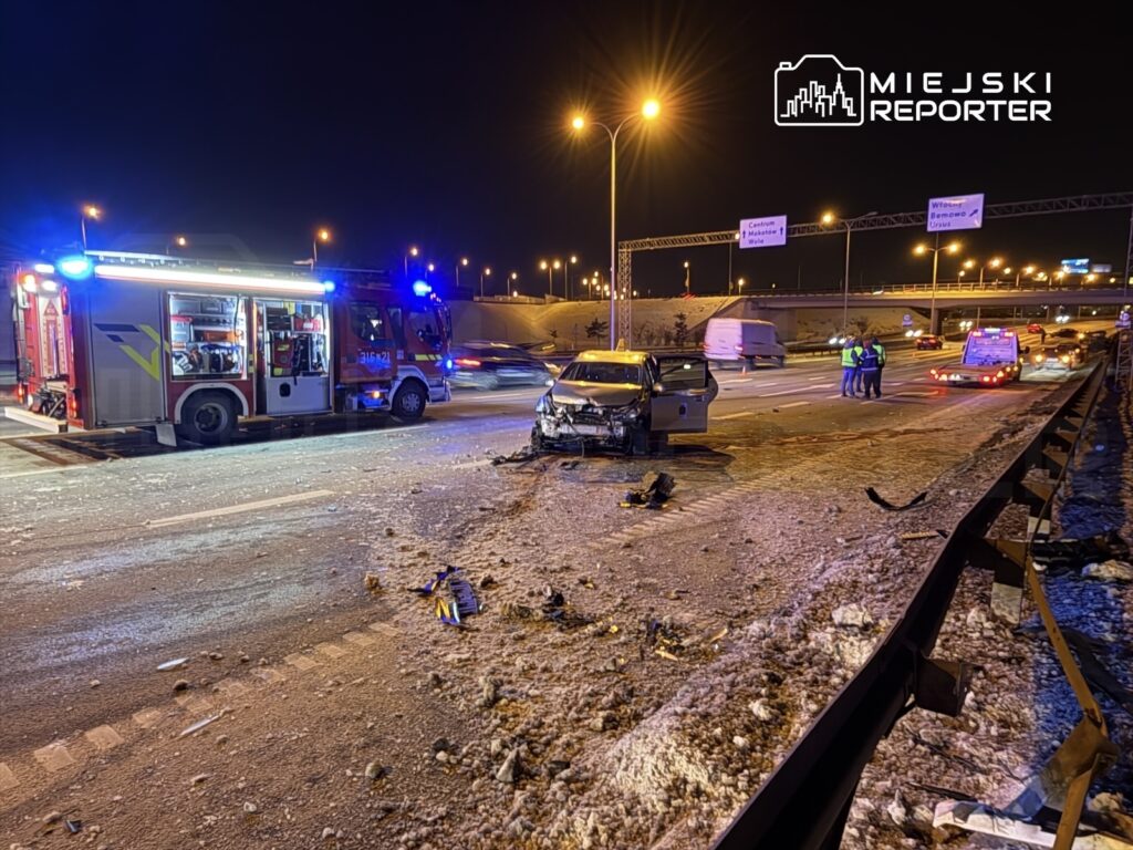 Uszkodzony samochód osobowy na drodze ekspresowej, obok stacja straży pożarnej z włączonymi sygnałami świetlnymi.