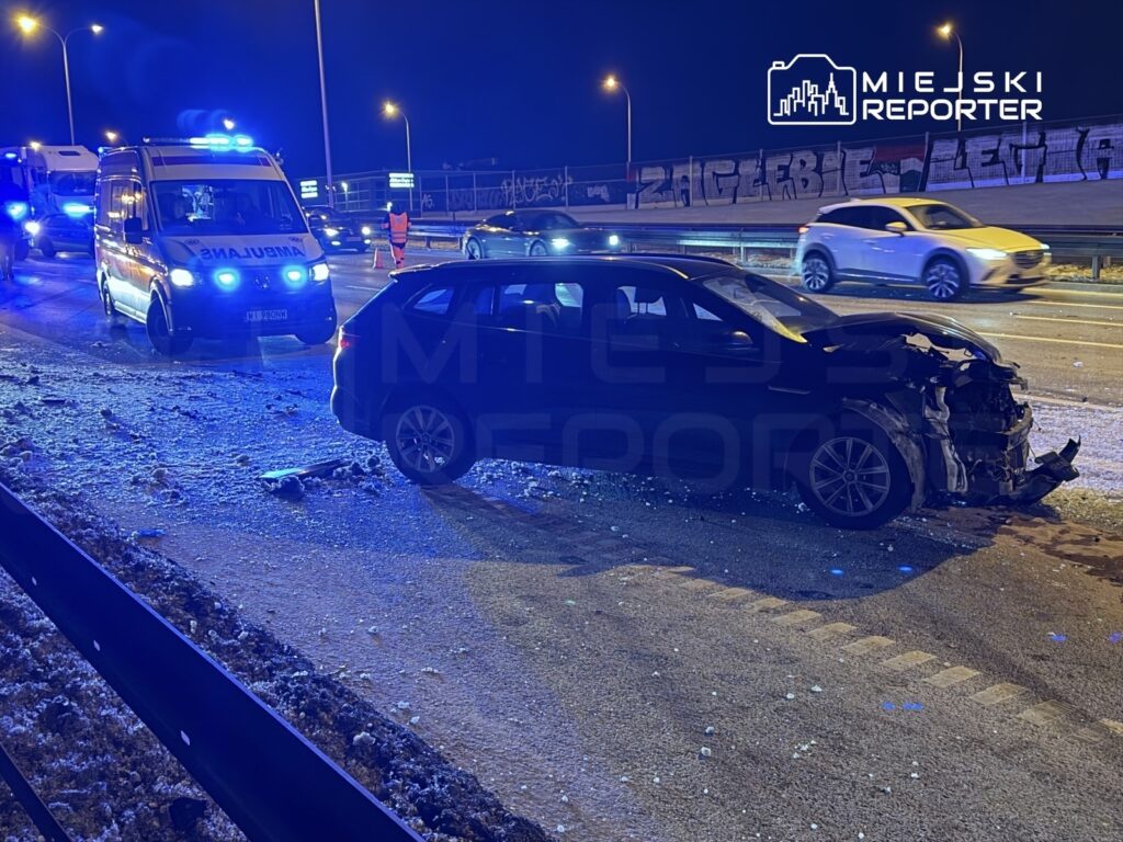 Czarny samochód osobowy z uszkodzoną przednią częścią stoi na poboczu drogi, obok niebieska karetka pogotowia.