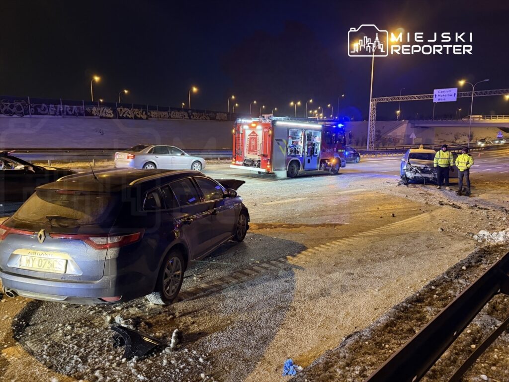Dwa uszkodzone samochody po kolizji na drodze, obok wozu strażackiego i dwóch funkcjonariuszy w odblaskowych kamizelkach.