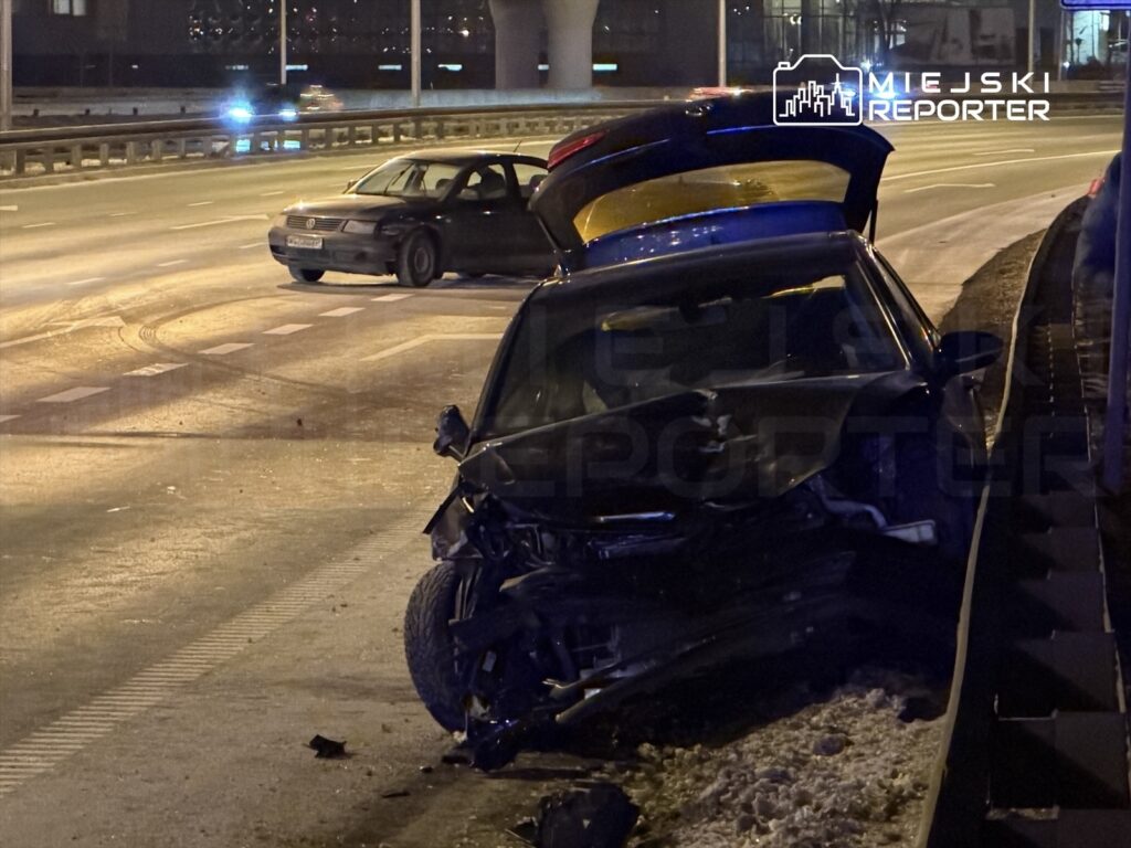 Zniszczony samochód osobowy z otwartą maską stoi na poboczu drogi, obok widać drugi pojazd. Nocna sceneria.