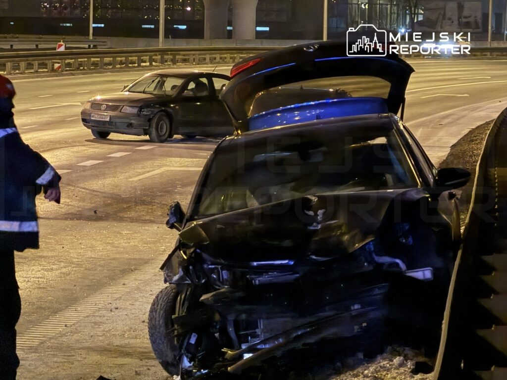 Strażak zbliża się do uszkodzonego czarnego samochodu na drodze, obok stoi inny pojazd.