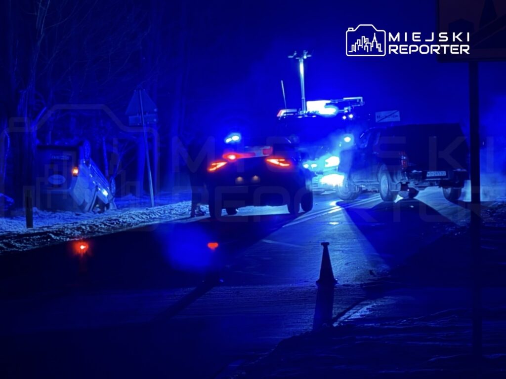 Niebieskie światła radiowozów oświetlają drogę, gdzie przewrócony samochód leży w rowie obok.