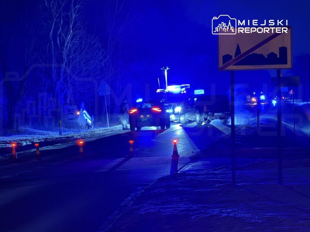 Na drodze w nocy stoją policyjne pojazdy, a w tle widać przewrócony samochód i migające niebieskie światła.