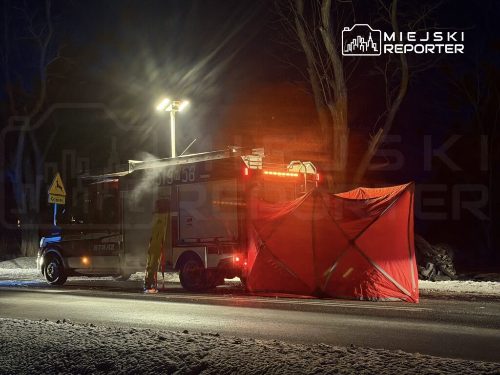 W nocy, w zimowym otoczeniu, widać wóz strażacki z włączonymi sygnałami świetlnymi, ustawiony obok czerwonego namiotu na drodze.
