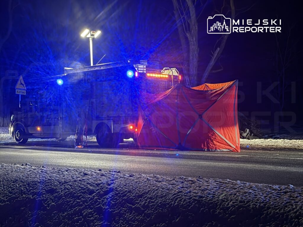 W nocy na drodze stoi wóz strażacki z włączonymi sygnałami świetlnymi, obok rozłożony pomarańczowy namiot.