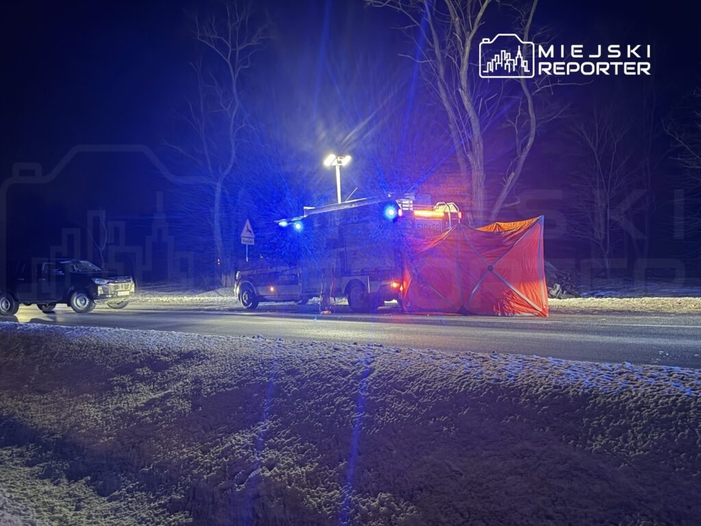 Pojazd policyjny z włączonymi sygnałami świetlnymi zabezpiecza miejsce zdarzenia na zaśnieżonej drodze nocą.