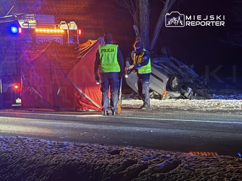 Policjanci w odblaskowych kamizelkach zabezpieczają miejsce wypadku, obok leży przewrócony samochód.
