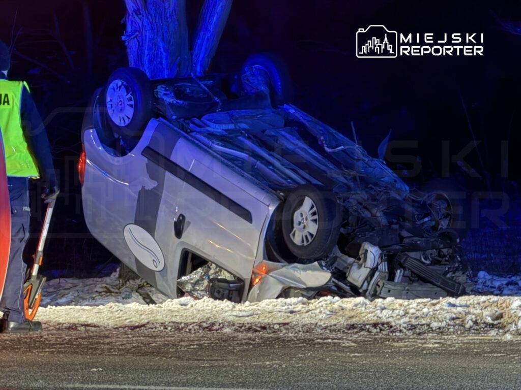 Policyjny funkcjonariusz z latarką sprawdza przewrócony pojazd na poboczu drogi w zimowej scenerii.
