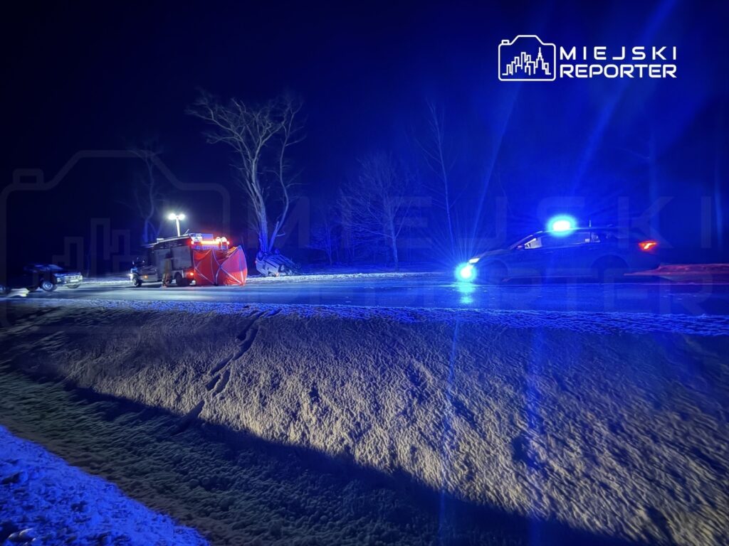 W nocy, na oświetlonej drodze, straż pożarna z czerwoną przyczepą oraz radiowóz z niebieskimi światłami uczestniczą w akcji.