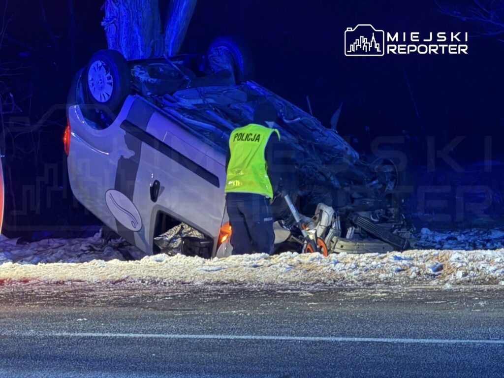 Policjant w kamizelce odblaskowej bada przewrócone na dach auto na poboczu drogi w zimowej scenerii.