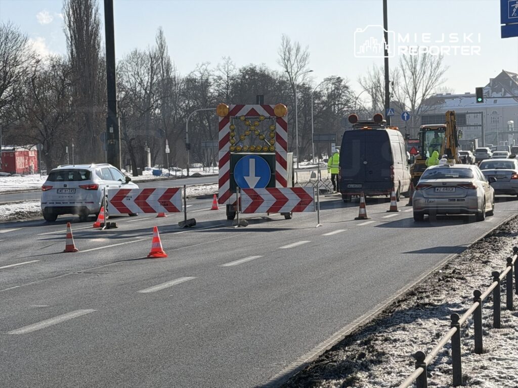 Pracownicy w odblaskowych kamizelkach prowadzą prace drogowe, a ruch jest kierowany przez znaki i pachołki.