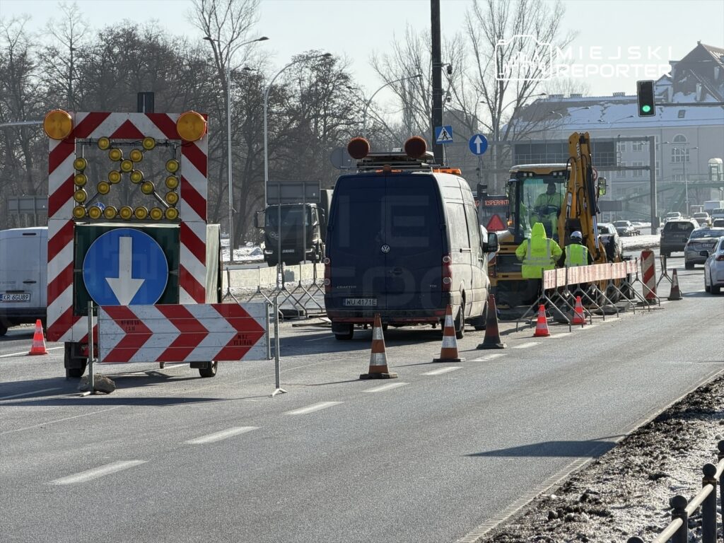 Robotnicy w żółtych kamizelkach pracują przy wykopie na jezdni, obok oznakowania drogi i pojazdów.