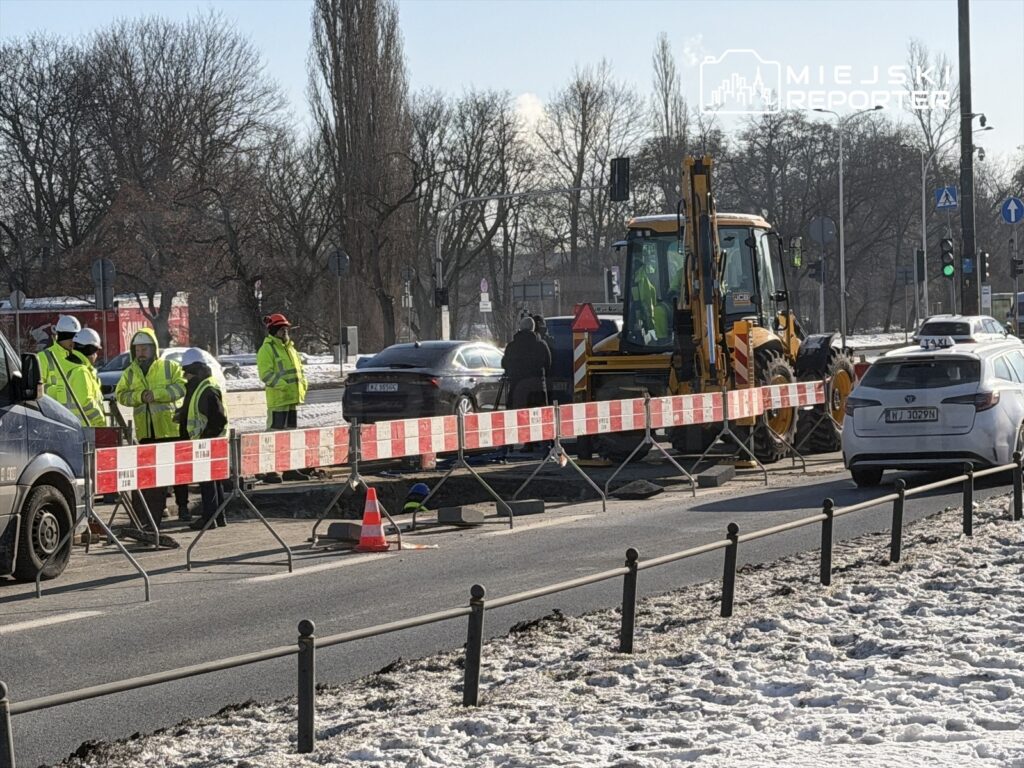 Grupa pracowników w odblaskowych kamizelkach prowadzi prace budowlane przy jezdni, obok znaku drogowym.