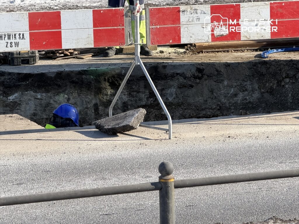 Robotnik w niebieskim kasku pracuje w wykopie drogowym, obok znajduje się barierka ochronna.