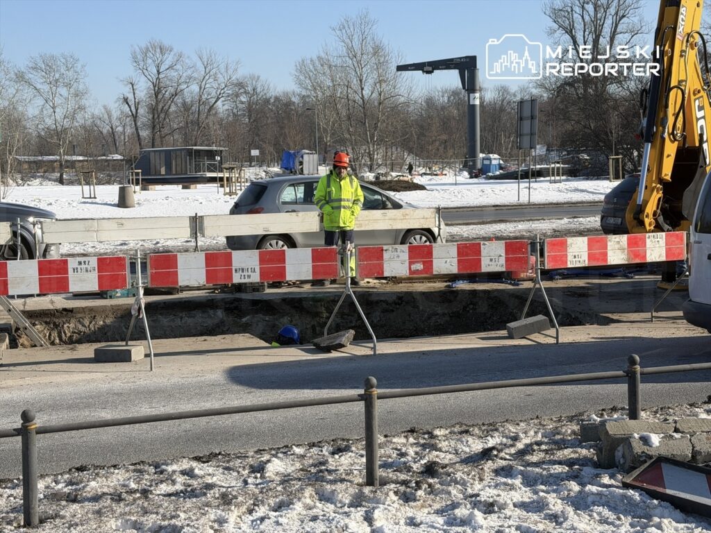 Robotnik w kamizelce odblaskowej nadzorujący prace budowlane przy otwartym wykopie na drodze, w zimowej scenerii.