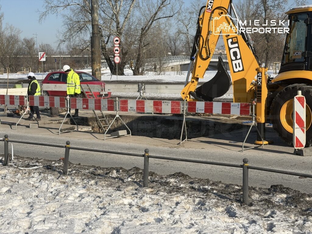 Dwaj pracownicy w odblaskowych kurtkach kontrolują wykop na drodze, obok stoi żółta koparka.