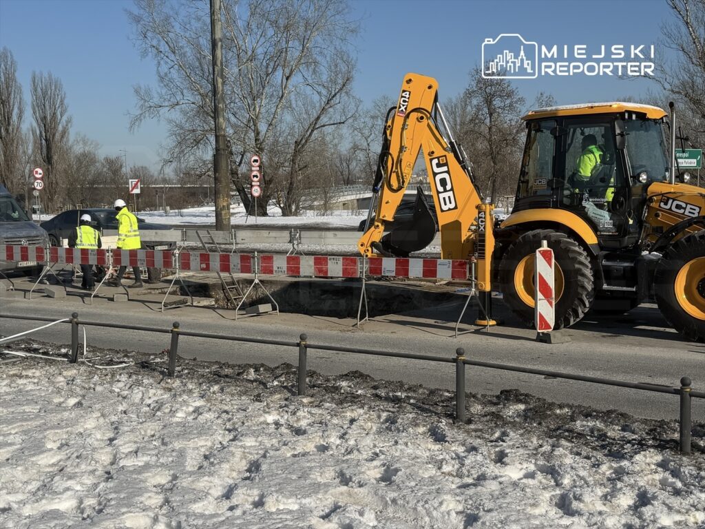Grupa pracowników w odblaskowych kamizelkach prowadzi prace drogowe obok koparki na śnieżnej drodze.
