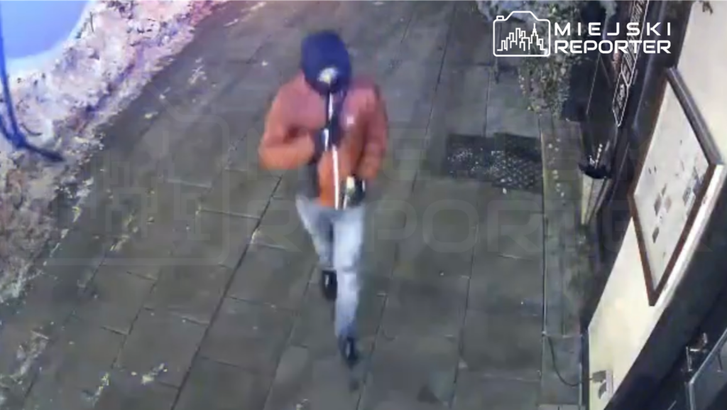 Mężczyzna w pomarańczowej kurtce idzie chodnikiem, trzymając w ręku przedmiot w pobliżu budynku.