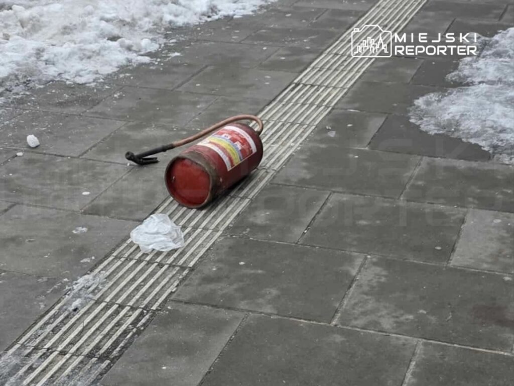 Na chodniku leży przewrócony gaśnica, obok niej znajduje się kawałek białego plastiku, a w tle widoczny śnieg.