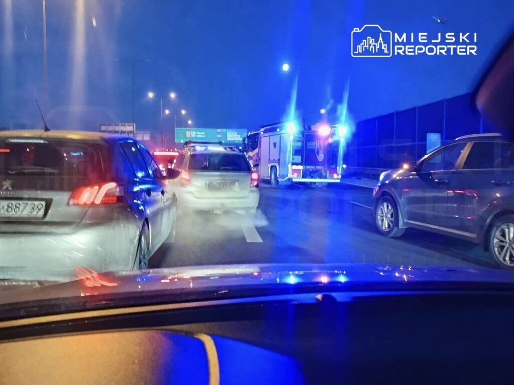 Ruch samochodowy spowolniony na drodze, w tle widać samochód ratunkowy z włączonymi sygnałami świetlnymi.