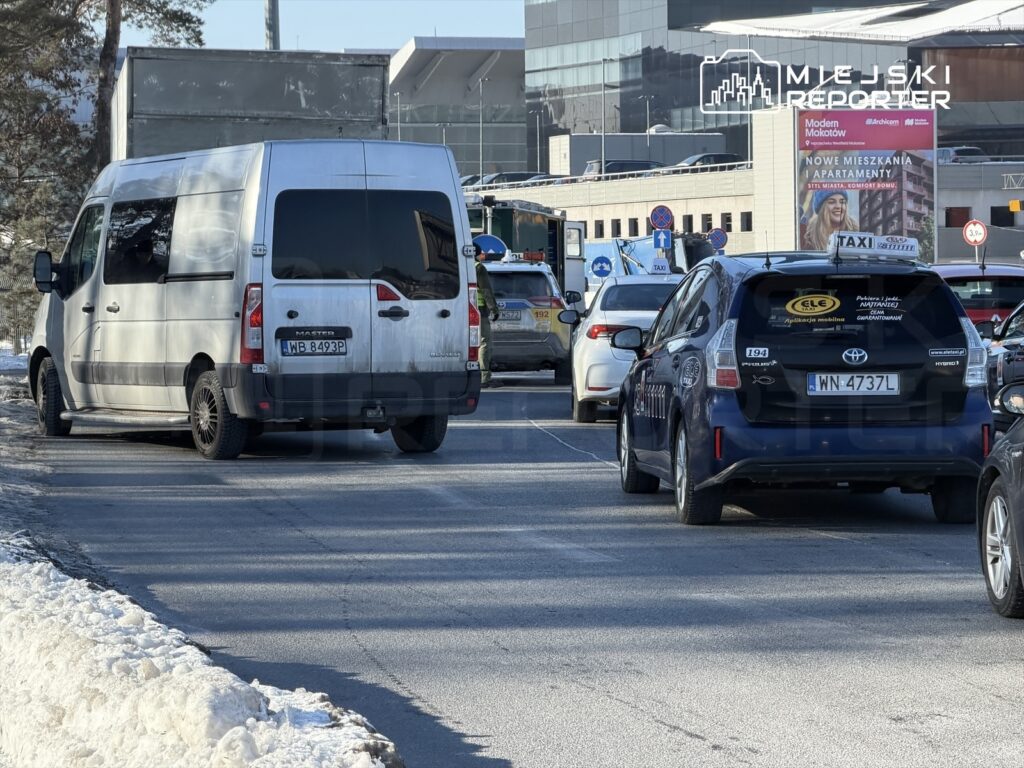 Biały bus dostawczy parkuje na ulicy obok taksówki w ruchliwym mieście, w tle widoczny nowoczesny budynek.