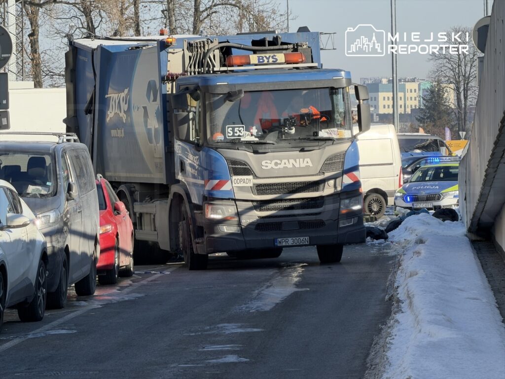 Śmieciarka marki Scania zatrzymana na wąskiej ulicy, obok zaparkowane samochody osobowe, w tle widoczne radiowozy policyjne.