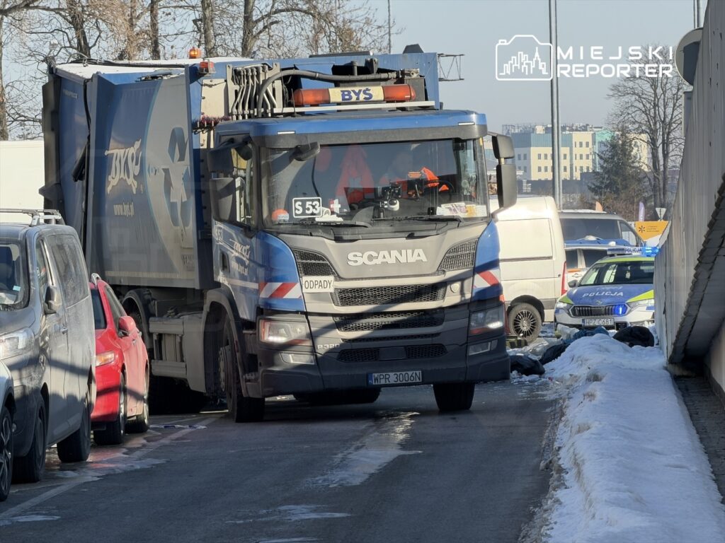 Śmieciarka marki Scania parkuje na ulicy, obok zaparkowane samochody, w tle widoczna policja i budynki.