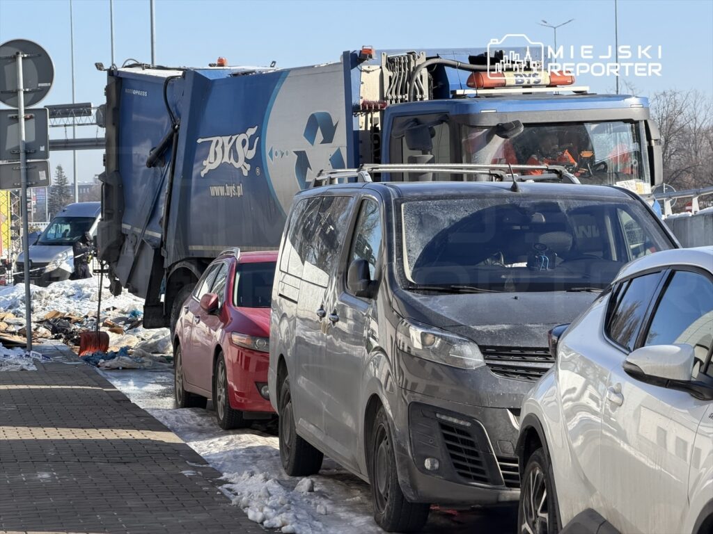 Śmieciarka z logo firmy zajmującej się recyklingiem zatrzymała się przy zaparkowanych samochodach na ośnieżonej ulicy.