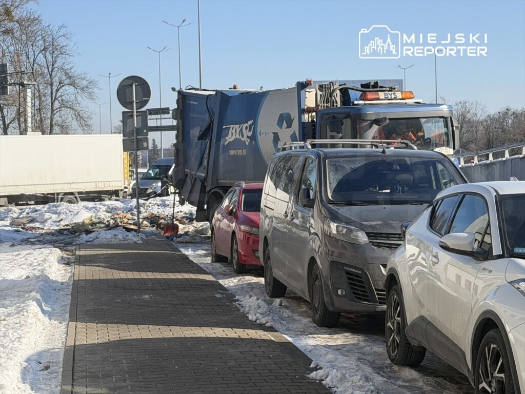 Śmieciarka zbierająca odpady na zaśnieżonej ulicy, obok zaparkowane samochody osobowe.
