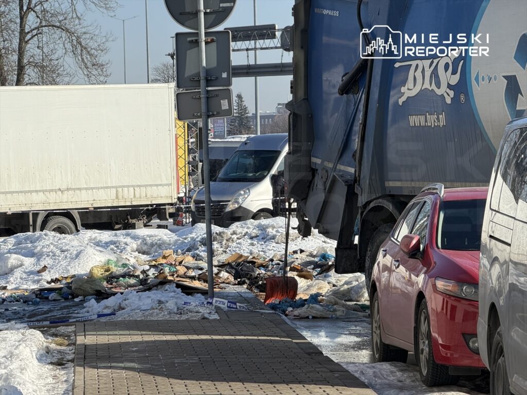 Na chodniku widać zanieczyszczenia, w tym odpady i śmieci, obok zaparkowane samochody oraz tiry w tle.