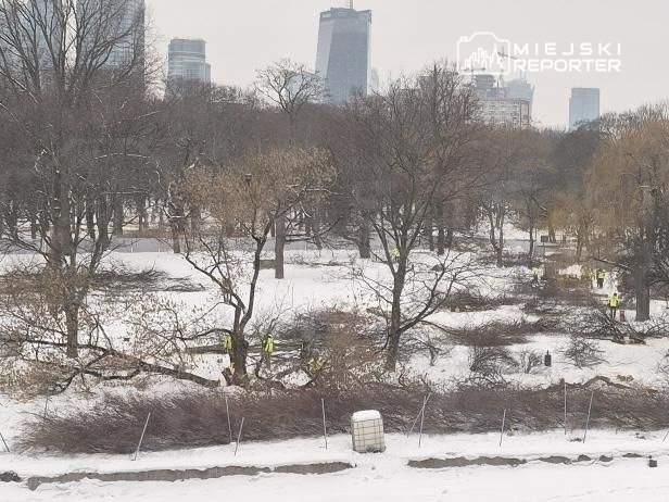 Pracownicy w żółtych kamizelkach porządkują teren parku pokrytego śniegiem, w tle nowoczesne budynki.