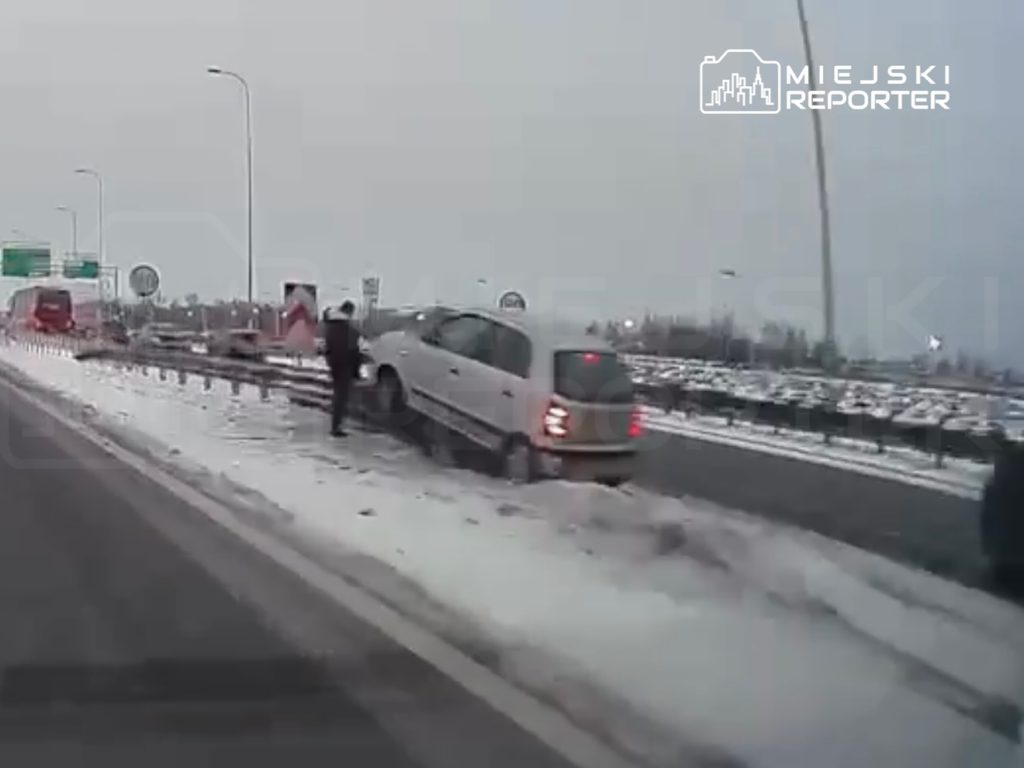 Kierowca samochodu osobowego wjeżdża na barierkę na ośnieżonej drodze, obok zatrzymane inne pojazdy.