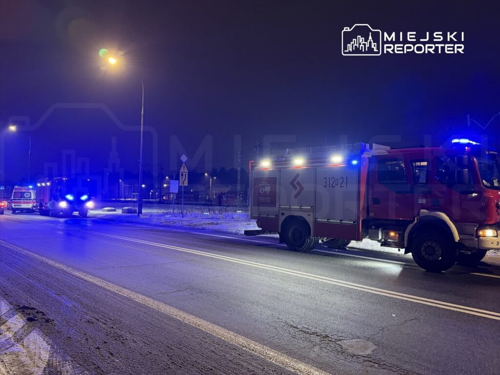 W nocy pojazdy straży pożarnej i karetka stoją na oświetlonej ulicy, emitując niebieskie światła.