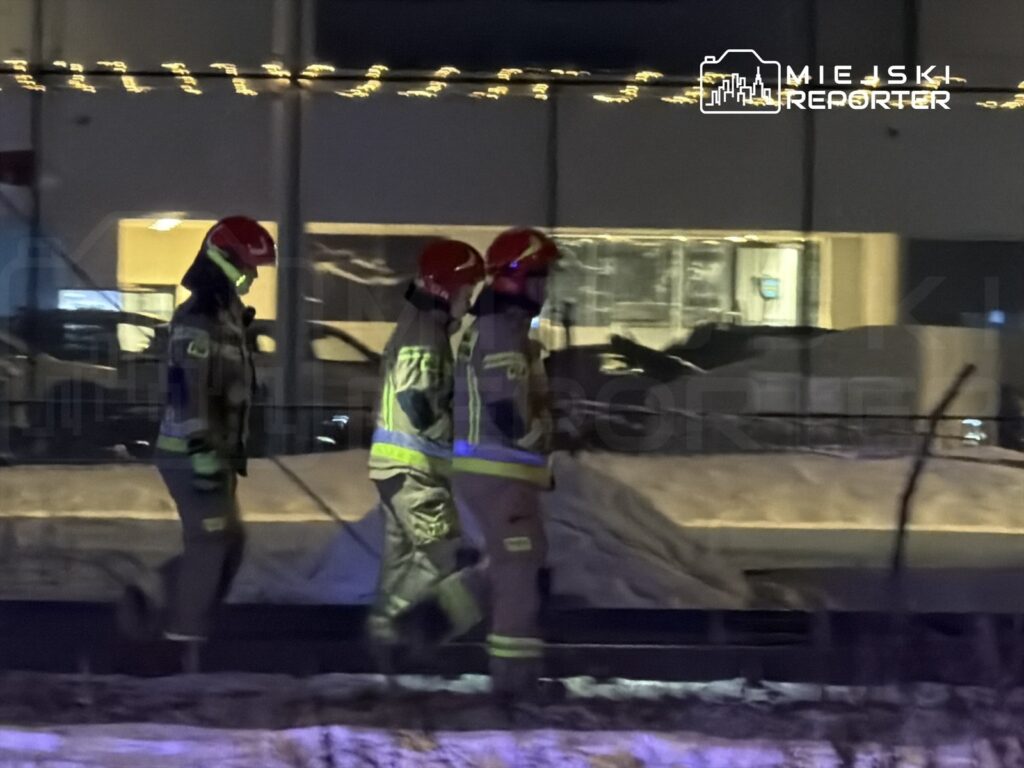 Grupa strażaków w hełmach przechodzi obok budynku, w tle widoczne oświetlenie świąteczne.