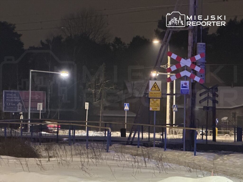 Latarnia oświetlająca przejazd kolejowy, przy którym znajduje się znak ostrzegawczy oraz billboard z informacją o stacji kontroli pojazdów.