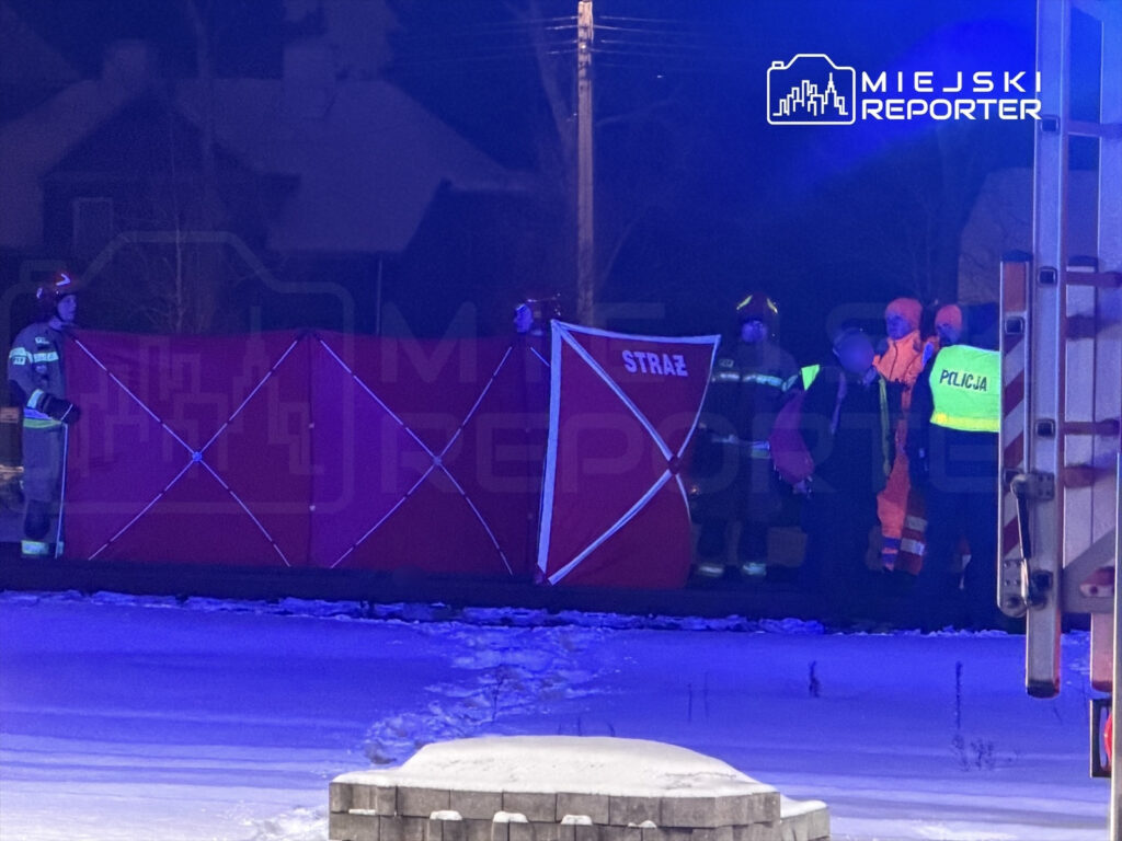 Grupa strażaków i policjantów pracuje przy rozstawionym czerwonym namiocie ochronnym w zaśnieżonym terenie nocą.