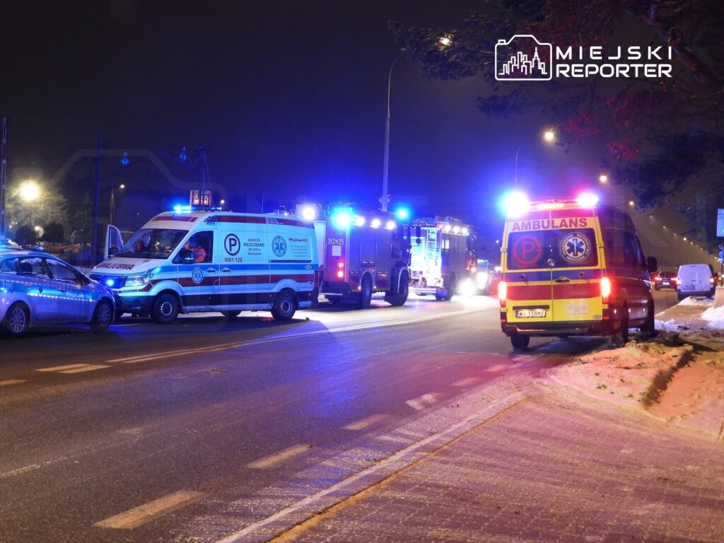 Ambulans oraz radiowóz policyjny zatrzymane na drodze, otoczone pojazdami ratunkowymi, w nocy z włączonymi sygnałami świetlnymi.