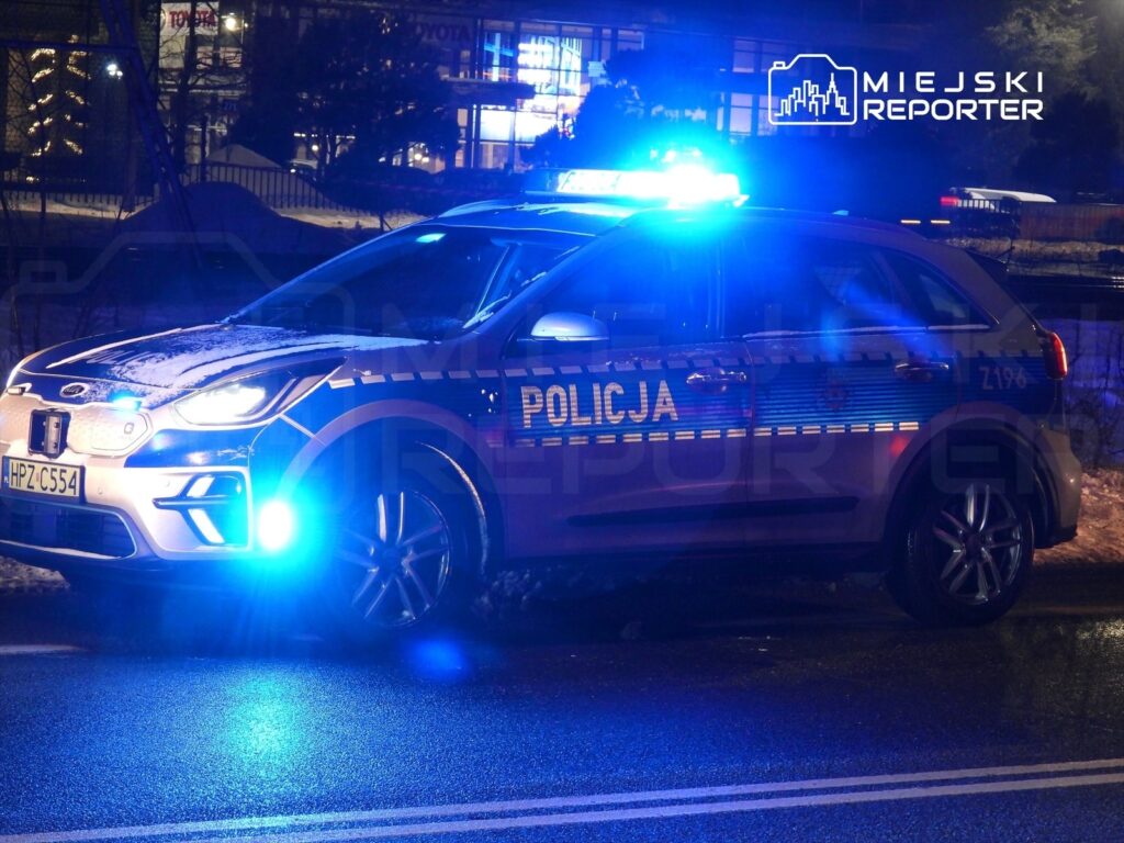 Policyjny samochód z niebieskimi światłami stoi na ulicy w nocnym otoczeniu, obok oświetlonych budynków.